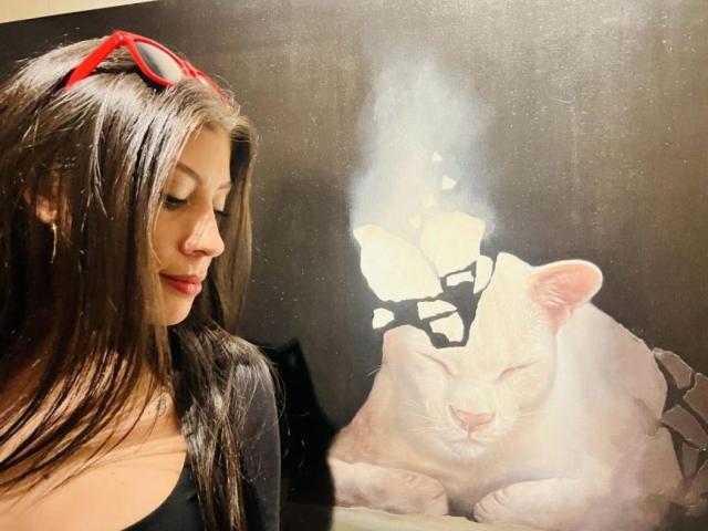 LizzieRossa with long brown hair, red sunglasses on head, black top, looking down at a white cat with closed eyes, lying on a dark surface.