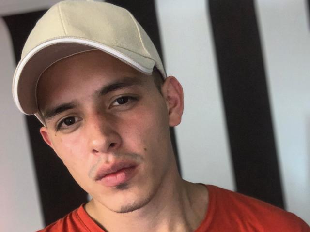 LiamEnzo with a light beige cap, red shirt, and a slight smirk, showing a small moles on his left cheek, with a neutral expression. His ass is not visible.
