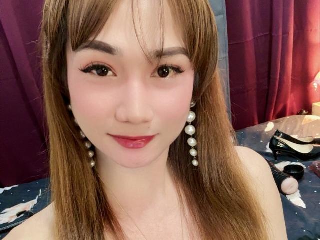AsianXPixyCumLust tiene el pelo largo y castaño, aretes de perlas y lápiz labial rojo. Está sonriendo, lleva un piercing en la nariz, mostrando su cara linda y un maquillaje sutil. No se ven los senos..