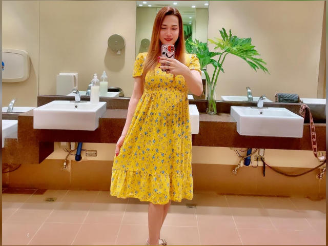 AsianXPixyCumLust vestindo um vestido floral amarelo, segurando um celular com um adesivo de rosto fofo, em pé em um banheiro, sorrindo, com uma planta verde ao fundo..