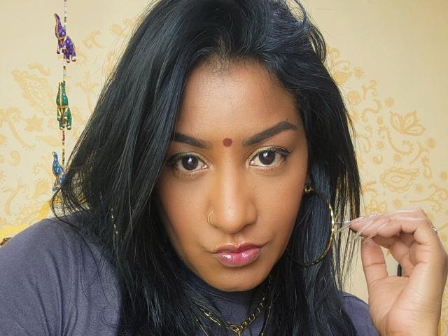 Nahirr has dark brown skin, black hair, brown eyes, a red bindi, gold hoop earrings, and a nose ring. She's wearing a purple top and looking serious.