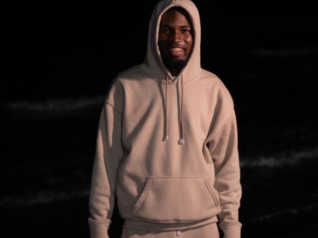 TysonKarter, dark-skinned, bearded, smiling, wearing a beige hoodie with a hood, standing in a dark night setting, hands in pockets, soft light highlighting his face.