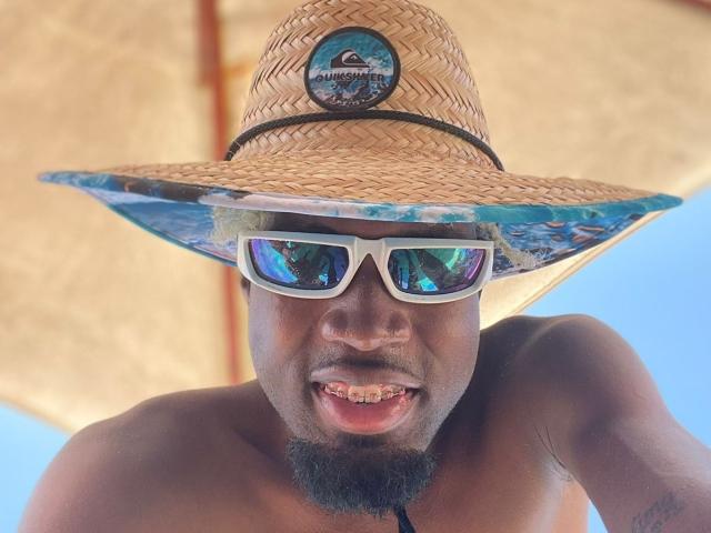TysonKarter, que lleva un sombrero de paja con un logotipo, gafas de sol reflectantes y no lleva camisa, muestra los dientes con una ligera sonrisa, tiene la piel oscura y un bigote..