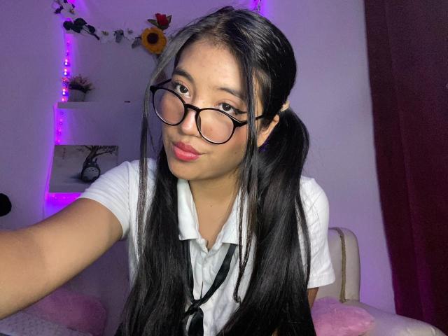 Elizaa, com maria-chiquinhas pretas, óculos e batom rosa, vestindo um uniforme escolar branco, tirando uma selfie, com os seios mal visíveis, sorrindo suavemente, em um quarto com pouca luz..