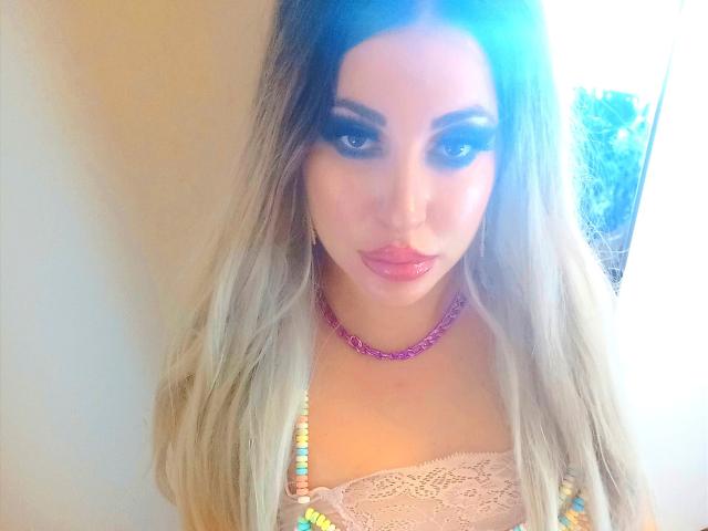 MissCha69 with long blonde hair, wearing a pink lace top, purple necklace, and colorful beads, looking at the camera with slightly pouted lips and dark eyeliner.