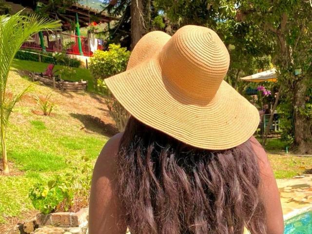 NahirobyEbony's back, wearing a big, tan, ribbed sunhat, with wavy, dark brown hair, standing by a pool, in a sunny, green, garden-like setting.