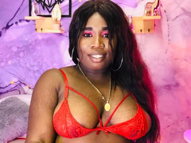 NahirobyEbony with dark skin, long black hair, pink eye makeup, red lace bra showing her big tits, gold necklace, smiling, wearing hoop earrings, in a purple-lit room.