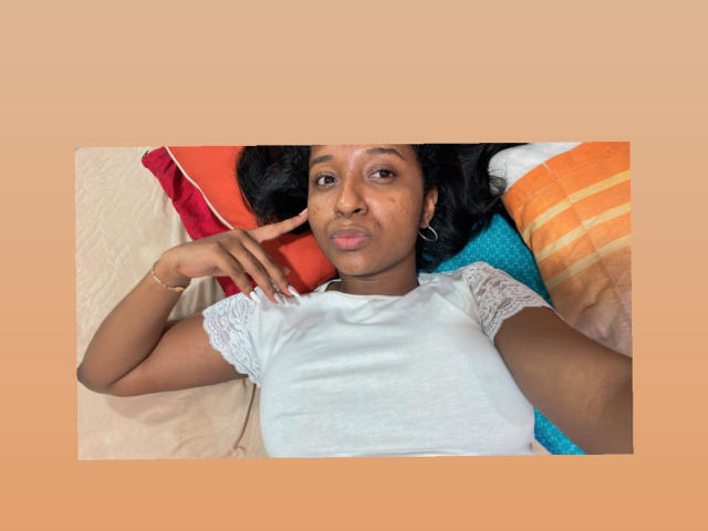 MelanyScoth lying on a bed, wearing a white lace-trimmed shirt, black hair, brown skin, small hoop earrings, pouty lips, hand near face, colorful pillows behind her.