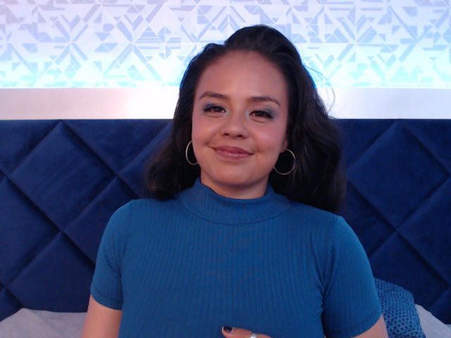 ArielleWilde with dark wavy hair, wearing a blue ribbed t-shirt, smiling at the camera, has small hoop earrings, and is holding her tits with her right hand.