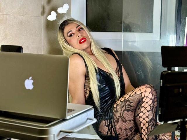 ShopieBurningT with long blonde hair, red lipstick, black lace bodysuit, and fishnet stockings, sitting on desk with laptops, looking sexy, showing ass, and a confident smile.