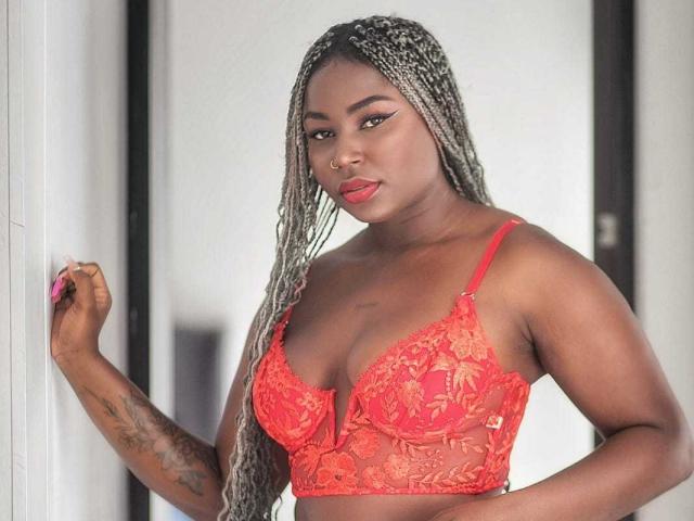 AkaneCox, with dark skin and long braided hair, wears a red lace bra, showing her tits and a tattoo on her right arm, standing against a white wall.