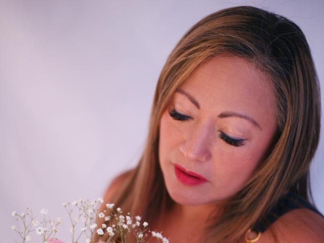 ChristinMoore with brown hair, looking down at small white flowers, has pink lipstick and black eyeliner, wearing a black top, her eyes are closed, in a soft light.