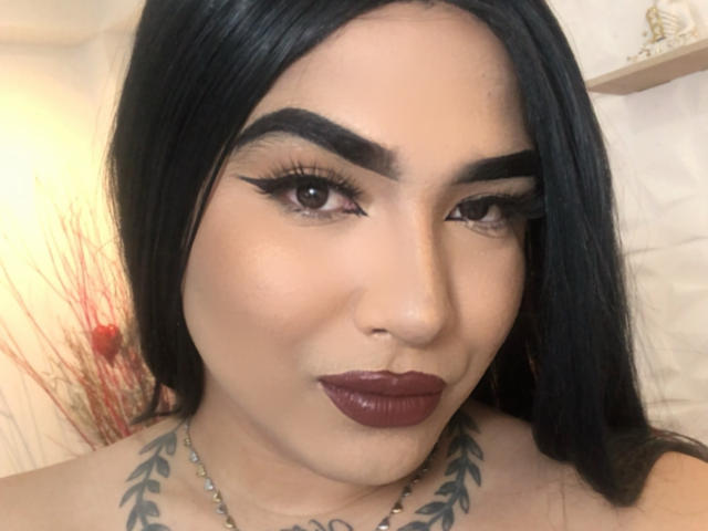 Emperatris has black hair, thick eyebrows, long eyelashes, dark red lipstick, and a tattoo with leafy patterns on her chest. Her skin is tan, and she's wearing a necklace.