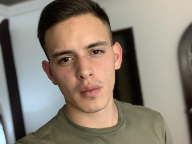 LiamEnzo has short brown hair, a light skin tone, and a neutral expression. He's wearing a green shirt, and his brown eyes are slightly downcast.