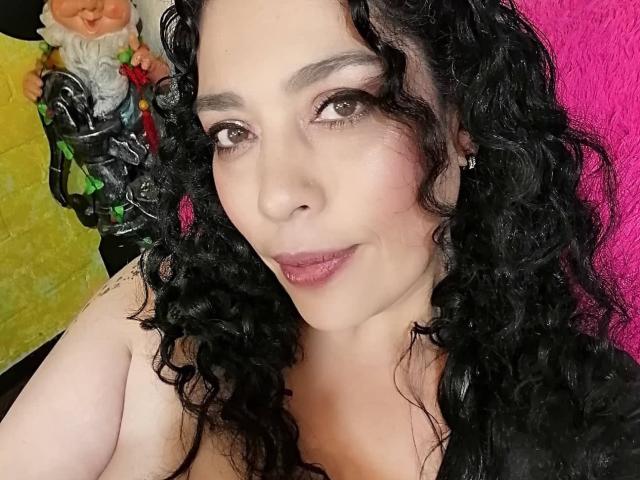 LyemDreams with curly black hair, brown eyes, and pink lipstick, showing off her tits, wearing a black top, against a pink background, with a gnome figurine on the left.