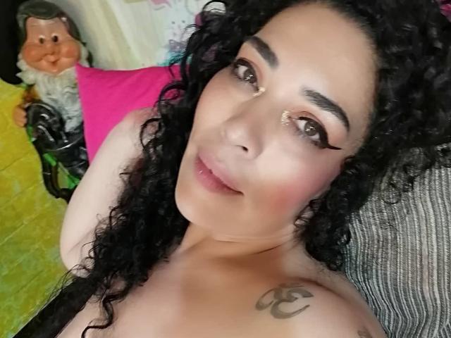 LyemDreams has curly black hair, brown eyes with long eyelashes, and a tattoo on her left shoulder. She's topless with pink and green pillows in the background.
