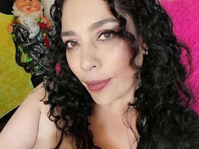 LyemDreams has curly black hair, brown eyes, and pink lipstick, with her tits partially visible, wearing black and pink in the background, and a gnome figure on the left.