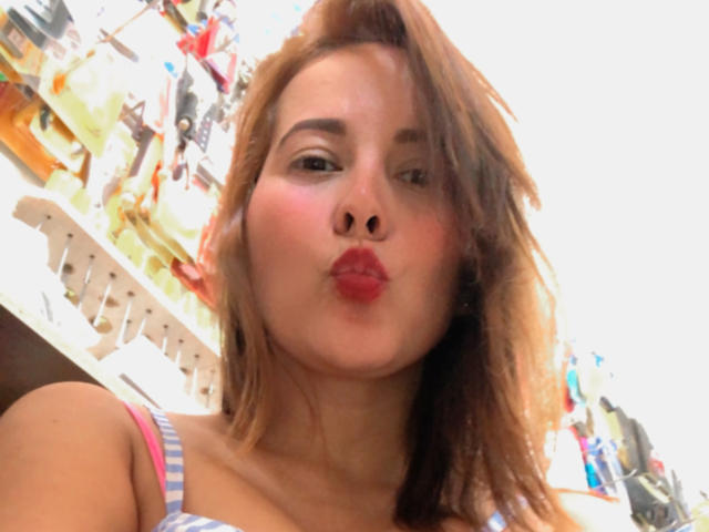 SaritaVelez, con labios rojos y sensuales, cabello ondulado castaño y una camiseta a rayas, besando la cámara, mostrando pequeños senos, piel clara y maquillaje sutil..