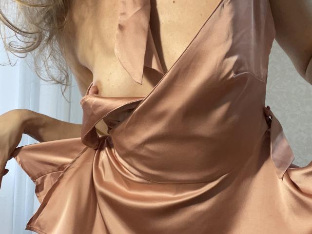 TheHottestBlonde's pink satin dress is pulled down, exposing her small tits with a ribbon. Her wavy blonde hair is visible, and her skin is light.