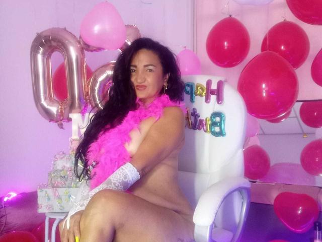 MarieStormy, naked, with long black hair, pink feather boa, white lace gloves, sitting on a white chair, surrounded by pink balloons and metallic "20" balloons, smiling at the camera.
