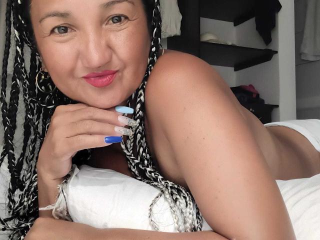 MarieStormy with braided black and white hair, pink lipstick, blue nail polish, lying on white pillow, smiling, showing her tits, hand near her chin, in a bedroom.