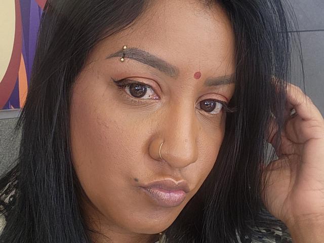 Nahirrs Gesicht mit dunklem Haar, braunen Augen, einem Nasenring und Make-up. Sie hat einen roten Punkt auf ihrer Stirn, goldene Piercings in den Augenbrauen und eine Sommersprosse in der Nähe ihres Mundes..