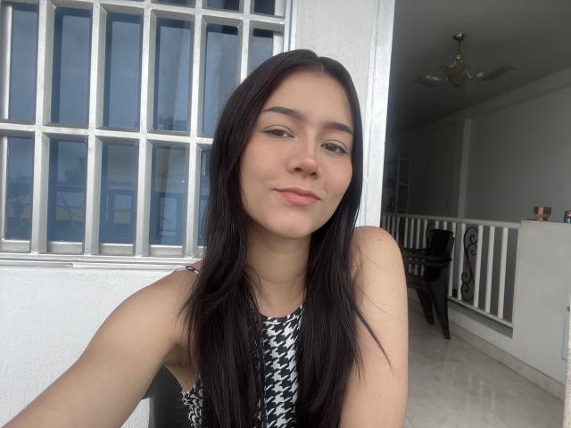 DeisyWest with long black hair, wearing a houndstooth sleeveless top, has a cute smile and is looking slightly to the side in a bright indoor setting.