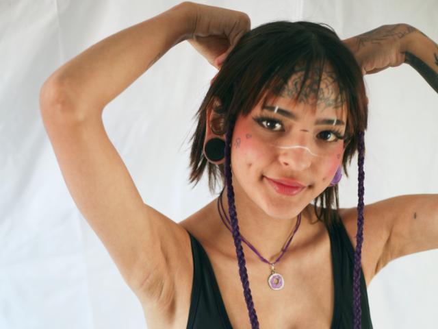 EmmaDemon with purple braids, black tank top, and purple necklace, arms raised, showing tattoos, large black ear gauges, and pink face paint, smiling at the camera.
