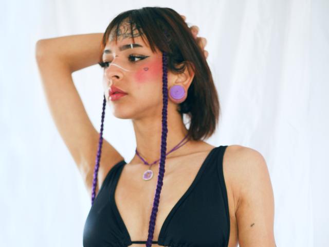EmmaDemon with purple braids, black dress, pink blush, purple earrings, and necklace, hand in hair, looking sideways, red lipstick, small tits, light skin, white curtain background.