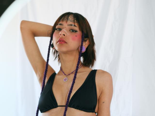 EmmaaDemon with purple braids, black bikini top, pink face paint, purple earrings, hand behind head, necklace, tattoo on forehead, slight smile, looking at camera.
