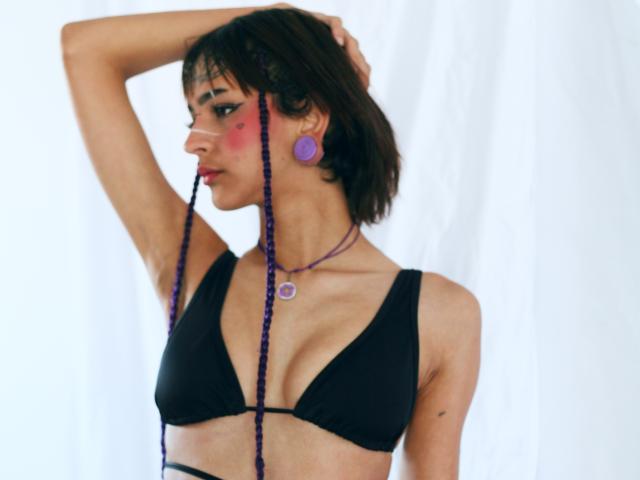 EmmaDemon with short brown hair, wearing a black bikini top, purple beads in mouth, pink blush, purple earrings, and purple necklace, hand in hair, looking side profile.