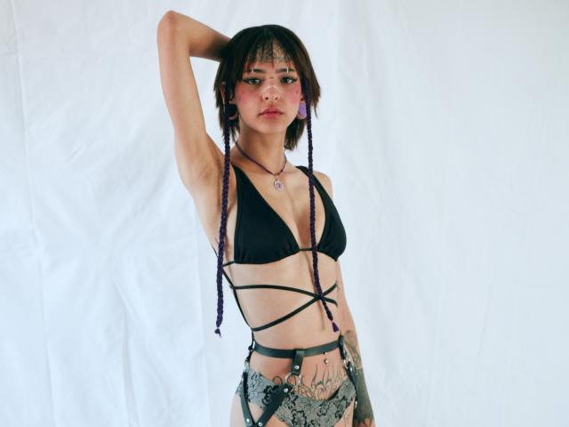 EmmaaDemon with short dark hair, purple braids, black strappy bra, tattoos, necklace, raised arm, looking at camera, white background, wearing lace bottoms, showing small tits, confident expression.