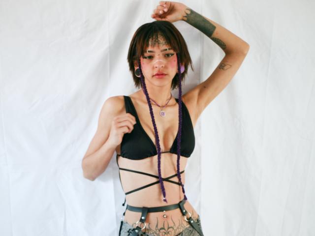 EmmaaDemon with purple braids, black strappy top, showing her tits, tattoos on arm and belly, white background, arm raised, looking at camera, wearing purple earrings.