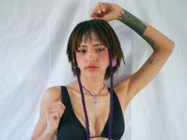EmmaaDemon with short brown hair, purple braids, black tank top, pink cheeks, tattoos, raised arm, purple earrings, purple necklace, looking down, white background, slight blur.