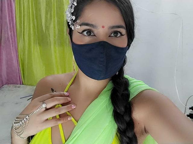 UmaRany, con cabello oscuro trenzado, lleva una máscara azul marino, un sari verde y un adorno de flores en el cabello, mostrando sus senos con una mano en el pecho..