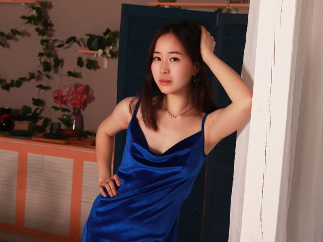 GinnyBean in a blue velvet dress, hand in hair, standing against a white wall, blue shutters in background, looking off-camera, small tits, light skin, black hair, subtle makeup.