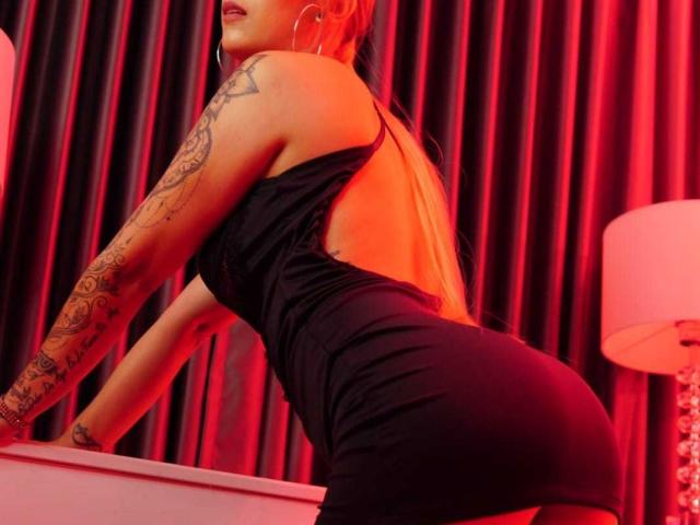 AnnieLaurenn in a tight black dress, leaning forward with her ass up, showing tattooed arms and red lighting, wearing hoop earrings, looking sexy and confident.