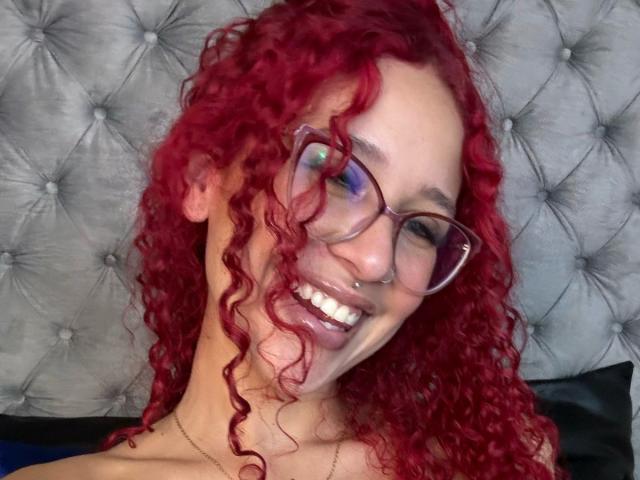 ValeryScot with curly red hair, wearing glasses, smiling, showing nose ring, and white teeth, against a tufted gray headboard, looking cute and slightly nerdy.