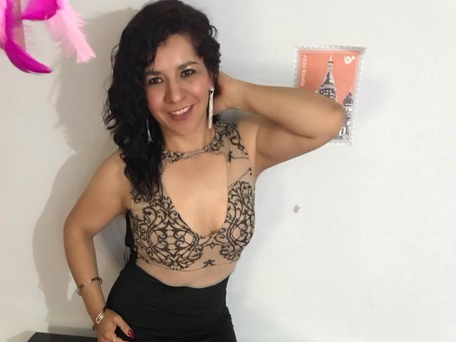EtoileLatin, con cabello negro rizado, sonriendo, vestida con una blusa marrón de encaje con cuello en V pronunciado y una falda negra, tiene tatuajes en el pecho y está de pie con una mano en la cabeza..