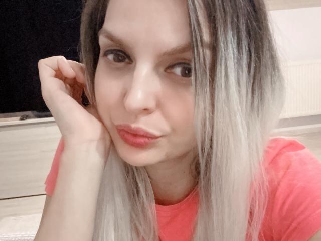 DouceAmelia with long, straight, blonde-to-gray ombre hair, pouty pink lips, and a cute nose, wearing a pink shirt, resting her chin on her hand.