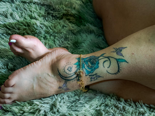 LyemDreams' foot with a blue rose tattoo, curly black lines, and small leaves, wearing a gold ankle bracelet, on a green fuzzy rug. Her white-painted toenail is visible.