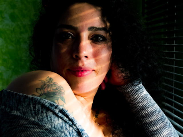 LyemDreams with curly black hair, pink lipstick, and gray off-shoulder sweater, showing a rose tattoo on her shoulder, smiling with sunbeam on her face.