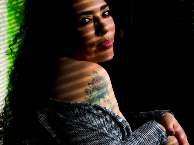LyemDreams with curly black hair, dark skin, and pink lipstick, showing a rose tattoo on her shoulder, in a grey off-shoulder top, with shadows on her face.