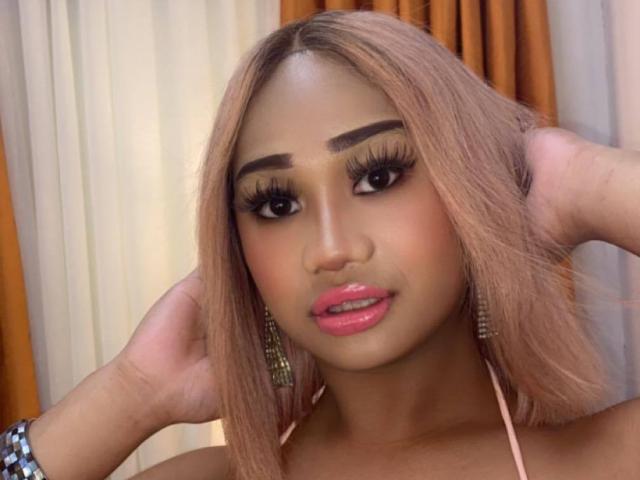 GiganticCummerx with pink hair, dark eyebrows, long eyelashes, pink lips, tan skin, and hands in hair, wearing pink halter and green earrings, looking at camera.