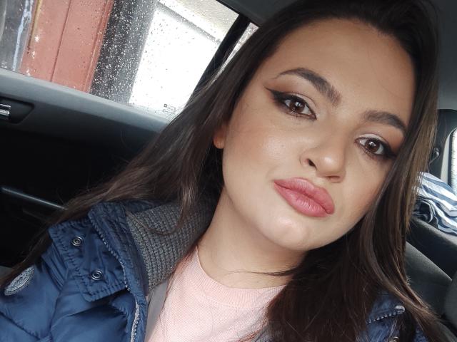 Raysaa tiene ojos marrones, labios rosados y el pelo largo y castaño oscuro, lleva una chaqueta azul sobre una camiseta rosa, y está sentada en un coche con lluvia en la ventana..