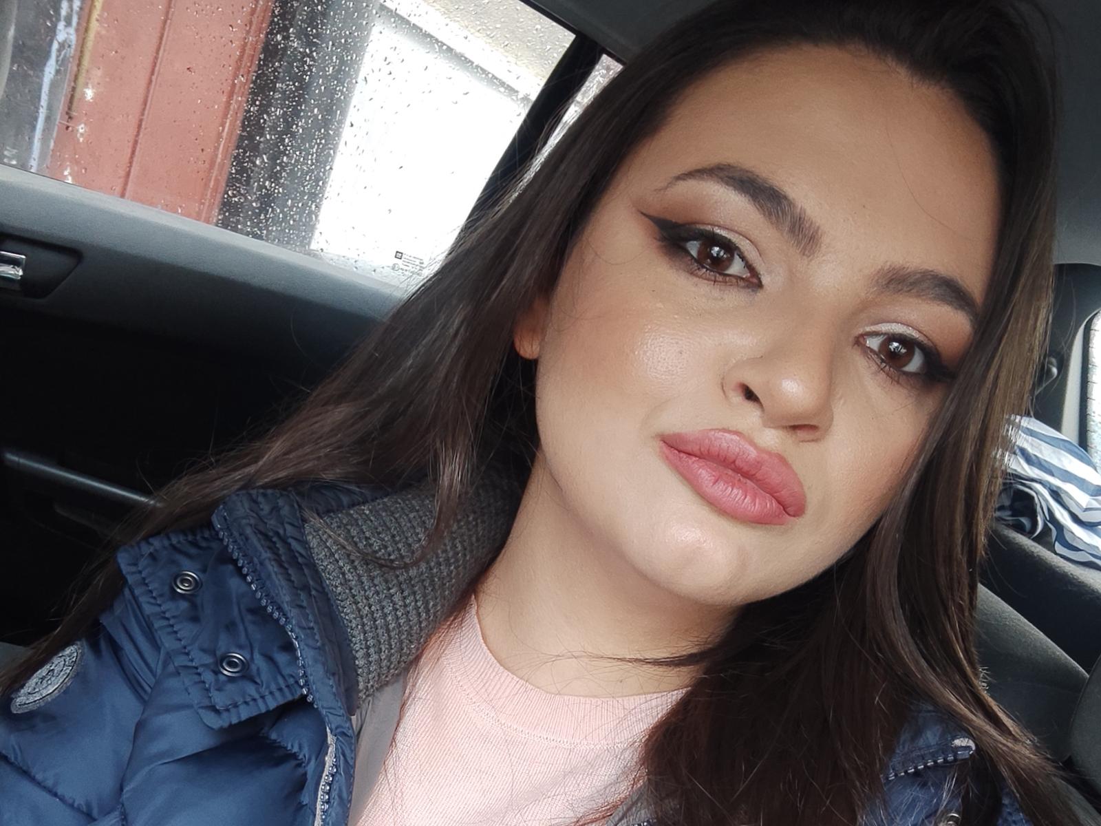 Raysaa has brown eyes, full pink lips, and long dark brown hair, wearing a blue jacket over a pink top, sitting in a car with rain on the window.