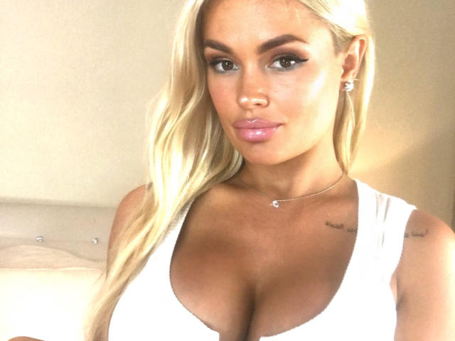 MilenaBiaa with long blonde hair, white tank top, showing her big tits, pink glossy lips, tattoo on right shoulder, necklace, earrings, and a confident, sexy look in a selfie.