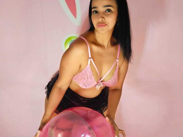 Amber Rousell mit schwarzen Haaren, einem pinken Spitzen-BH und einem schwarzen Rock, die nach vorne gebeugt ist, mit den Händen auf einem pinken Luftballon, lächelnd, wobei ihre Brüste sichtbar sind, vor einem pinken Hintergrund..