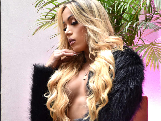 LorentsTS with long blonde wavy hair, black furry coat, and nude top, showing her tits, hand on chin, looking down, in front of a green plant.
