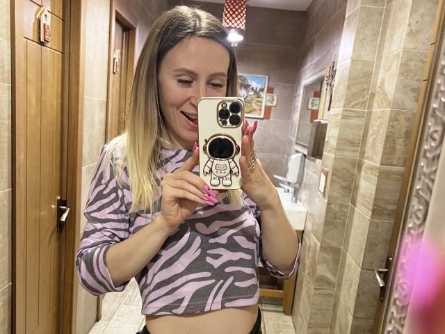 Saveta, con i capelli biondi, indossa un top corto a righe zebrate, sorride, tiene in mano un telefono con un pop socket, mostra il seno, ha le unghie rosa, e si sta scattando un selfie allo specchio del bagno..